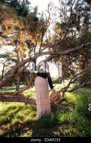 Une jeune femme enceinte se tient gracieusement dans une forêt ensoleillée, embrassant la beauté de la nature. Elle tient un chapeau, avec la lumière du soleil filtrant à travers les arbres, cr Banque D'Images