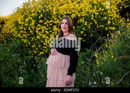 Une jeune femme enceinte se tient paisiblement devant des fleurs jaunes vibrantes, embrassant la beauté de la nature. Son expression calme reflète la sérénité Banque D'Images