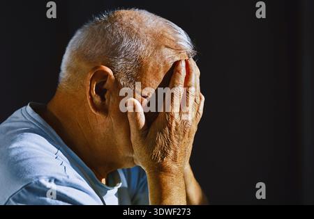 Un homme âgé aux cheveux gris est assis de profil avec ses mains obscurcissant son visage, capturant les détails et la texture de sa peau ridée, reflétant un Banque D'Images