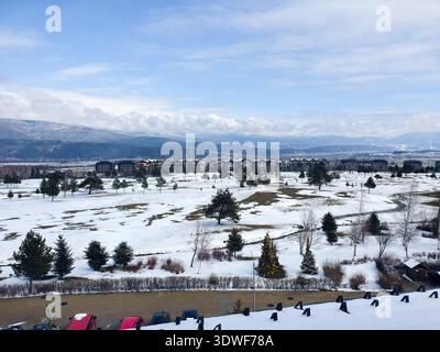 Terrain de golf enneigé et champs ouverts parsemés d'arbres à feuilles persistantes, encadrés par des bâtiments de villégiature éloignés et des montagnes vallonnées sous un ciel bleu vif Banque D'Images