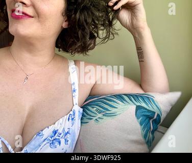 Femme souriante se relaxant à l'intérieur sur un canapé, portant une robe florale et tenant ses cheveux, portrait de style de vie décontracté. Banque D'Images