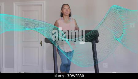 Femme senior marchant en haut rose, leggings bleus à l'aide d'un tapis roulant à la maison avec recouvrement de vague turquoise Banque D'Images