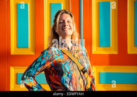 Une femme souriante se tient joyeusement devant une porte colorée dans le Salento, en Colombie. Sa chemise à motifs complète le dos orange et jaune éclatant Banque D'Images