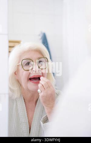 Une femme âgée avec des cheveux gris et des lunettes applique du rouge à lèvres rouge devant un miroir de salle de bain. Vêtue d'une robe légère, elle applique soigneusement le maquillage, Banque D'Images