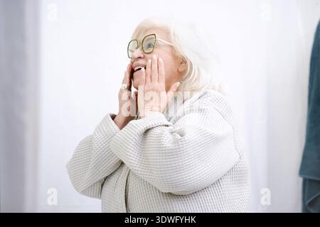 Femme âgée joyeuse dans des lunettes et une robe confortable sourit joyeusement, embrassant un moment de détente et de soins personnels à la maison, touchant son visage avec cont Banque D'Images