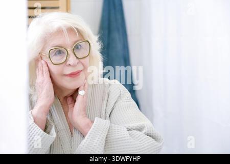 Une femme âgée dans une robe se tient dans une salle de bain lumineuse, réfléchissant paisiblement devant un miroir. Elle porte des lunettes et sourit doucement, dégageant un sens Banque D'Images