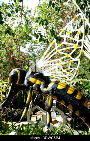 Vue rapprochée d'une sculpture d'abeille aux couleurs noires et jaunes et aux ailes complexes, sur fond de verdure luxuriante et de fleurs colorées Banque D'Images