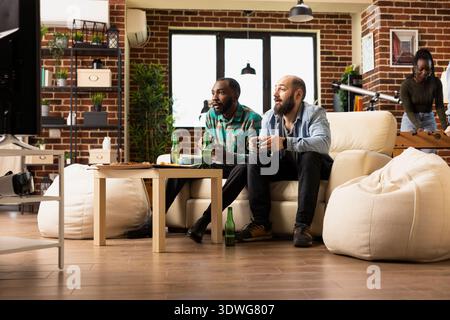 Hommes barbus multiethniques utilisant des manettes sans fil pour jouer à des jeux vidéo regardant l'écran de télévision dans la surprise. Jeunes amis masculins traînant et se liant sur le jeu multijoueur en ligne. Banque D'Images