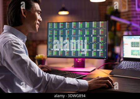 Heureux entrepreneur masculin assis avec un pc de bureau et un ordinateur portable, analysant des graphiques financiers. Homme d'affaires focalisé travaille à domicile, étudiant les investissements et les statistiques de trading numérique sur plusieurs écrans. Banque D'Images