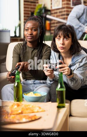 Gros plan sur diverses femmes tenant des manettes de jeu, axées sur une session de jeu multijoueur intense. Amis féminins se liant sur des jeux vidéo, des collations et des boissons dans un appartement de mur de briques confortable. Banque D'Images