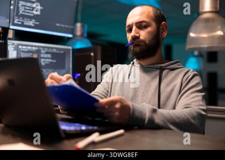 Le programmeur concentré feuillette les documents, recoupe les informations sur l'ordinateur portable. Le développeur masculin adulte tient le stylo et examine soigneusement les données sur le presse-papiers, garantissant ainsi la précision dans l'interface technique. Banque D'Images