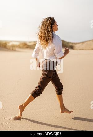 Jeune femme en chemise blanche courir pieds nus à travers la dune de plage de sable. Liberté, énergie et concept de mouvement en bord de mer Banque D'Images