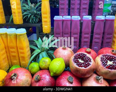Une variété de fruits frais tels que les grenades, les citrons et les ananas disposés avec des bouteilles de jus colorées dans un environnement de marché animé Banque D'Images