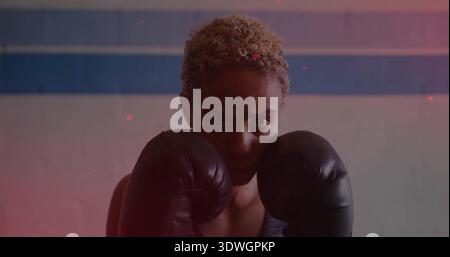 Tenant boxer féminin soulevant des gants noirs près du visage dans la salle de gym, portant un débardeur foncé, fusées roses Banque D'Images