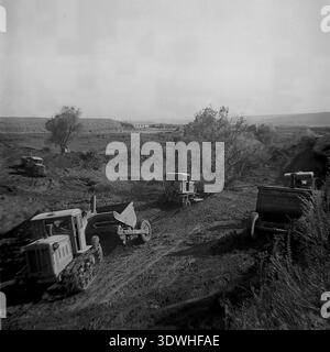 La photo historique en noir et blanc des années 1970 capture une flotte de lourds tracteurs à chenilles soviétiques tirant des racleurs pendant les travaux de terrassement à Donrybkombinat dans le district de Slavyansk, RSS d'Ukraine, URSS. Opérant sur un vaste paysage de terre battue, les machines construisent et agrandissent des étangs artificiels pour l'aquaculture. Cette image d'archives authentique met en évidence la mécanisation lourde, la remise en état des terres à grande échelle et le développement constant de l'infrastructure agricole dans le Donbass pacifique avant la guerre, reflétant la croissance régionale. Banque D'Images
