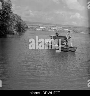 La photo historique en noir et blanc des années 1970 capture un bateau spécialisé motorisé d'alimentation de poissons sur un vaste étang à Donrybkombinat dans le district de Slavyansk, RSS d'Ukraine, URSS. Le navire, équipé d'une grande trémie pour distribuer la nourriture, se déplace à travers l'eau calme. Cette image d'archives authentique met en évidence la culture intensive des poissons d'étang, la mécanisation de l'aquaculture soviétique et le développement constant de l'industrie alimentaire dans le Donbass pacifique avant la guerre. Banque D'Images