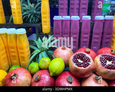 Une variété de fruits frais tels que les grenades, les citrons et les ananas disposés avec des bouteilles de jus colorées dans un environnement de marché animé Banque D'Images