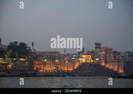 Ghats illuminés avec des bateaux sur le fleuve Gange pendant la soirée à Varanasi, en Inde, mettant en valeur l'architecture historique et la vie fluviale. Banque D'Images