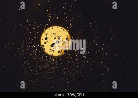 Gouttes de pluie et de lumière de la lune sur verre foncé la nuit. Verre de fenêtre humide avec beaucoup de gouttes de pluie et une vue sur le ciel noir de nuit avec le jaune Banque D'Images