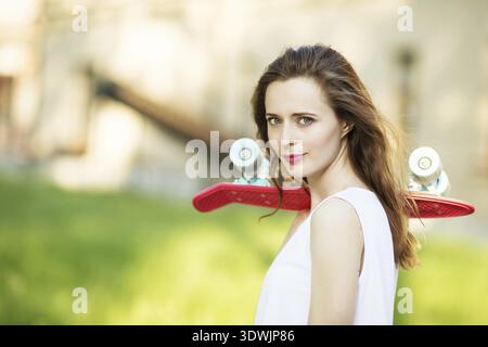 Portrait de belle fille urbaine en robe blanche avec un skateboard rose. Heureuse femme souriante. Fille tenant une planche à roulettes en plastique à l'extérieur. Vie urbaine Banque D'Images