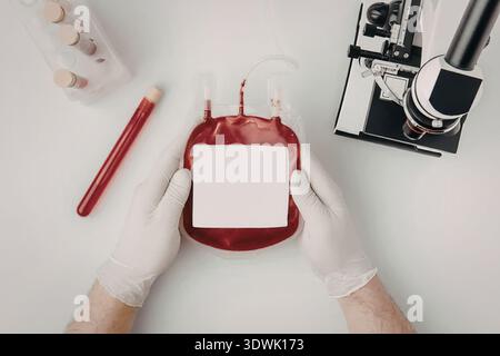 Une main gantée tenant une poche de sang avec une étiquette blanche à côté de l'équipement médical sur une surface blanche. Banque D'Images