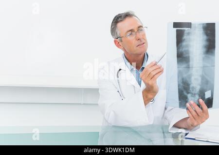 Médecin masculin senior assis sur un bureau tenant un stylo pointant le film de radiographie de la colonne vertébrale portant une blouse de laboratoire, espace de copie. Médecin, radiographie, consultation, clinique, guérison Banque D'Images
