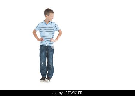 Garçon d'âge scolaire posant les mains sur les hanches regardant à gauche souriant dans un polo blanc et un Jean studio, espace copie. Portrait, jeunesse, vêtements décontractés, denim, chaussures, lig Banque D'Images