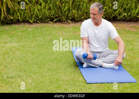 Homme senior assis sur un tapis d'exercice bleu sur la pelouse tenant un petit haltère bleu, espace de copie. Extérieur, jardin, arrière-cour, fitness, mobilité, force, plus vieux Banque D'Images