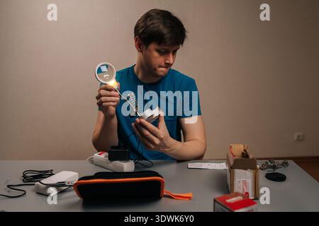 Homme se concentrant sur un appareil électronique, inspectant les composants avec une loupe éclairée tout en se préparant à la soudure, démontrant la précision dans une maison Banque D'Images