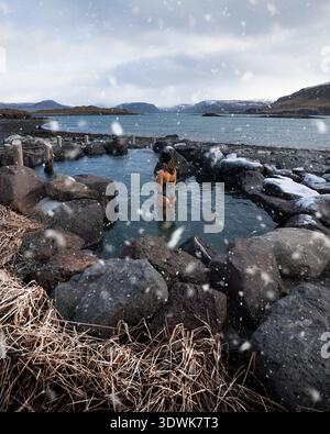 Vue d'une personne se dresse à la taille dans une piscine géothermique entourée de rochers et de neige, créant un contraste chaleureux avec le paysage froid, Mosfellsbae Banque D'Images