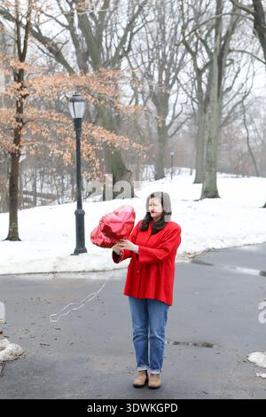 Femme en manteau rouge vif tient des ballons de coeur dans le parc enneigé de Brooklyn Banque D'Images