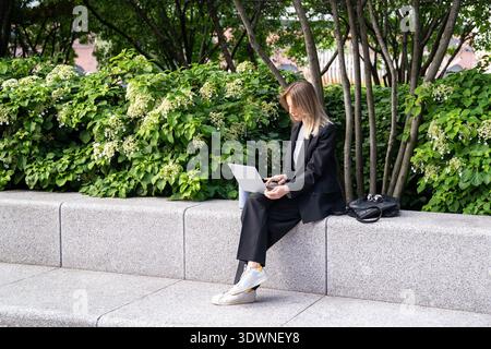 Femme d'affaires réussie travaillant à distance du bureau dans le parc de la ville en utilisant un ordinateur portable profitant d'une journée ensoleillée. Banque D'Images