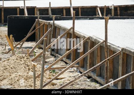 Les ouvriers préparent une zone pour une nouvelle dalle de béton avec une charpente en bois, des pieux de sable et des outils dispersés sur le sol sous la lumière du soleil. Banque D'Images