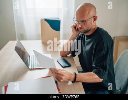 Homme chauve sérieux avec des lunettes travaillant intentionnellement dans le bureau à domicile lumineux, lisant la paperasse au bureau en bois avec ordinateur portable et smartphone, axé sur le professi Banque D'Images