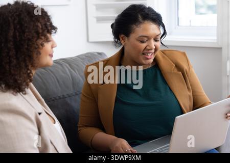 Des femmes afro-américaines collègues dans des blazers discutant du travail sur le canapé gris à l'aide d'un ordinateur portable argenté Banque D'Images