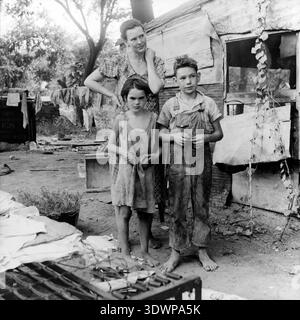 Cette poignante photographie en noir et blanc de Dorothea Lange de 1936 capture une mère et ses deux enfants pieds nus debout stoïquement devant leur maison de fortune en Oklahoma pendant la Grande dépression. L'image exprime avec force les difficultés et la résilience des familles déplacées par le Dust Bowl. Banque D'Images
