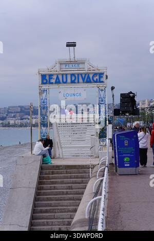 Arche d'entrée du salon plage beau Rivage sur la promenade de Nice, avec des marches vers la plage de galets, les gens, le drapeau français et la vue sur la mer Méditerranée couverte. Banque D'Images