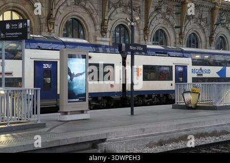 Le train régional bleu et blanc du Zou repose sur la plate-forme historique de la Gare de Nice ville sous de grandes arches en pierre et un panneau d'affichage numérique lumineux. Banque D'Images