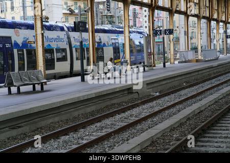 Moment calme à la Gare de Nice ville : un voyageur solitaire s'assoit sur un banc sous des colonnes dorées tandis qu'un train bleu Zou 'merveilles du Sud' repose Banque D'Images