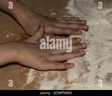 Mains d'enfant façonnant la pâte à pain fraîche sur une table en bois farinée. Un moment de cuisson maison chaleureux - parfait pour les campagnes de style de vie familial, paren Banque D'Images