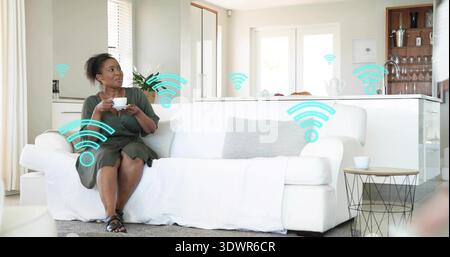Assise femme d'âge moyen tenant une tasse sur un canapé blanc, regardant à gauche à la maison, signaux wifi, espace de copie Banque D'Images