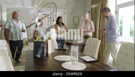 Amis riant debout autour d'une grande table à manger à la maison, versant du vin et montrant des sacs cadeaux Banque D'Images