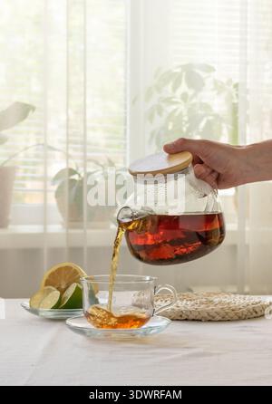 Une main verse le thé infusé de la théière en verre dans une tasse, avec des tranches de citron et des cubes de sucre sur la table près de la fenêtre Banque D'Images