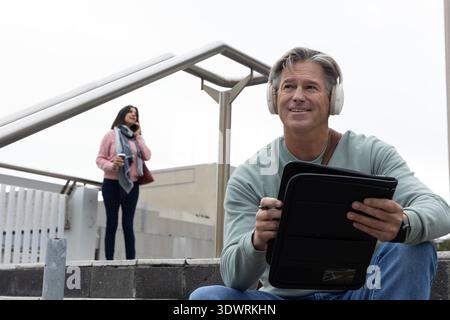 Homme et femme d'âge moyen assis et debout sur l'escalier urbain à l'aide d'une tablette avec stylet et téléphone Banque D'Images