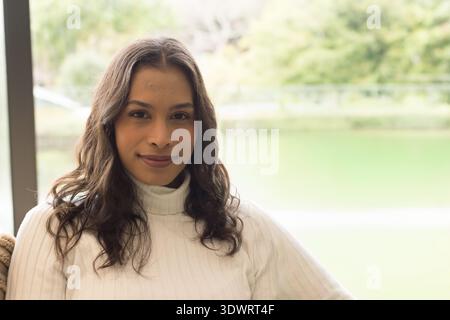 Femme assise par une grande fenêtre à la maison, portant un col roulé blanc, regardant l'étang et les arbres. Portrait, calme, serein, naturel, intérieur, doux, minimal Banque D'Images