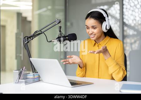 Jeune femme biraciale enregistrant un podcast, portant des écouteurs et parlant dans un microphone professionnel tout en regardant un ordinateur portable, partageant du contenu dans un espace de travail moderne Banque D'Images