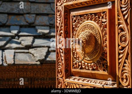 Gros plan d'une porte d'entrée en bois sculptée avec art au temple Narayan dans le village de Roghi avec un frappeur de porte en laiton détaillé et une toiture en pierre. Banque D'Images
