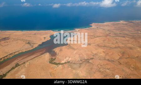 Vue aérienne d'un paysage côtier sec avec une rivière ou un estuaire se jetant dans l'océan bleu, terrain sablonneux rouge-brun, végétation clairsemée, ombres nuageuses Banque D'Images