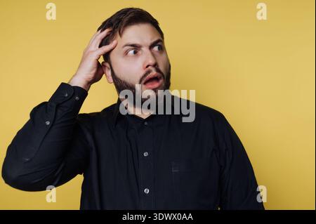 Un homme se tient face à un fond jaune vif, les yeux larges et la bouche agapée dans la surprise. Sa main repose sur sa tête, exprimant clairement un moment de choc Banque D'Images