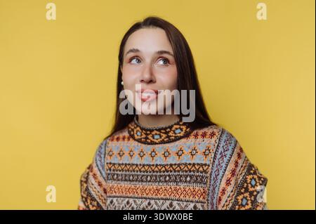 Une jeune femme aux cheveux longs et fluides partage un sourire chaleureux devant un fond jaune vif. Son pull coloré ajoute à l'atmosphère joyeuse, Banque D'Images
